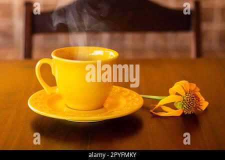 Goiania, Goias, Brésil – 01 mai 2023 : une tasse jaune remplie de café chaud, à la vapeur, avec une soucoupe et un tournesol, sur une table en bois et à l'arrière de Banque D'Images