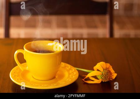 Goiania, Goias, Brésil – 01 mai 2023 : une tasse jaune remplie de café chaud, à la vapeur, avec une soucoupe et un tournesol, sur une table en bois et à l'arrière de Banque D'Images