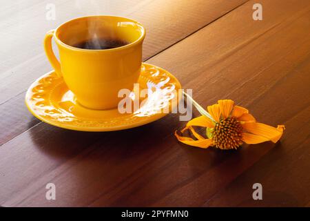 Goias, Brésil – 01 mai 2023 : une tasse jaune remplie de café chaud, en train de sortir, avec une soucoupe et un tournesol, sur une table en bois. Banque D'Images