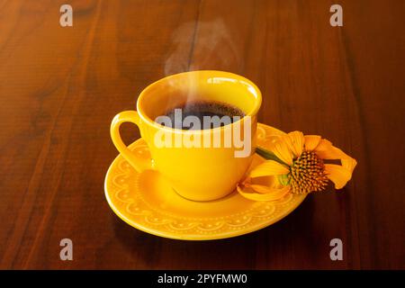 Goias, Brésil – 01 mai 2023 : une tasse jaune remplie de café chaud, en train de sortir, avec une soucoupe et un tournesol, sur une table en bois. Banque D'Images