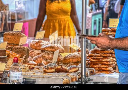 Boulangerie sur le marché avec l'accent sur le premier plan - vendeur vérifiant le téléphone comme femme dans la robe jaune approche - rogné et méconnaissable Banque D'Images