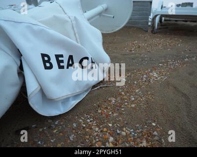 Un parasol était plié sur une plage de sable fin après le mauvais temps. Tempête en mer. Fin de la saison des plages en raison du typhon et du cyclone. Chaises longues, parasols et mobilier d'hôtel. Mot de plage écrit sur blanc Banque D'Images