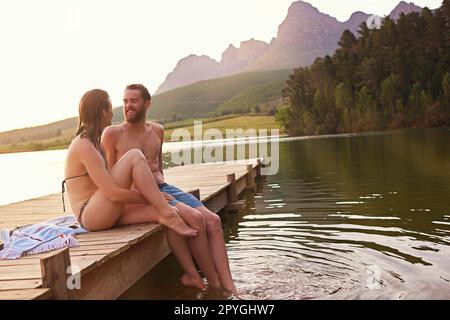 Été idyllique. un jeune couple affectueux en maillots de bain assis sur un quai au coucher du soleil. Banque D'Images