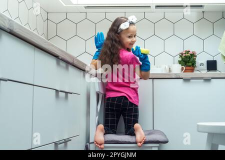 Heureuse petite fille dans des gants de protection laver la vaisselle avec une éponge dans l'évier de cuisine assis sur la chaise. Nettoyage à domicile. Banque D'Images