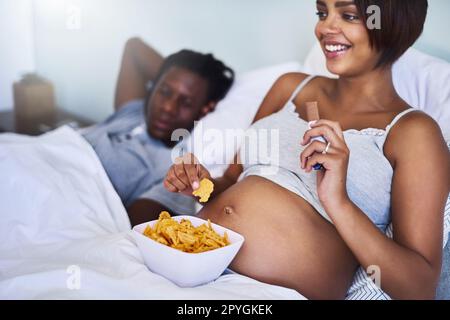 Je m'installe avec des en-cas heureux. une femme enceinte qui profite d'un chocolat et de pommes de terre au lit. Banque D'Images