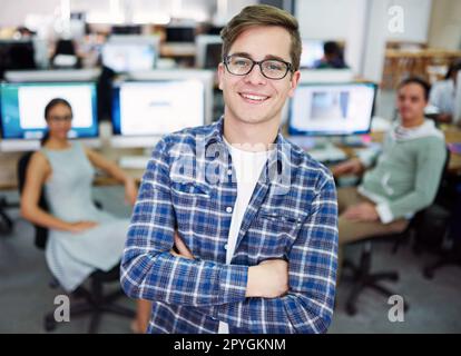 HES un jeune pro talentueux. Portrait d'un jeune homme debout dans un bureau avec des designers en arrière-plan. Banque D'Images