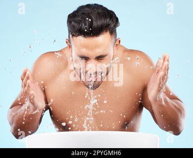 Visage, éclaboussures d'eau et soins de la peau de l'homme nettoyant en studio isolé sur un fond bleu. Hygiène, gouttes d'eau et lavage de modèle masculin, bain ou toilettage pour une peau saine, le bien-être du visage ou la beauté. Banque D'Images