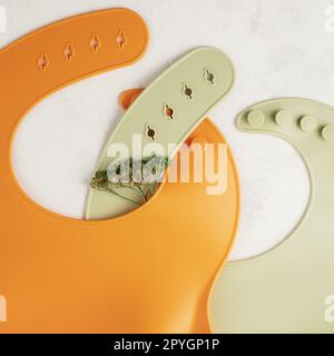 Deux bavoirs en silicone orange et vert croisés avec fleur verte séchée sur fond blanc. Accessoires pour bébé, concept d'alimentation Banque D'Images