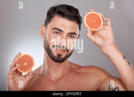 Portrait de visage, soins de la peau et homme avec pamplemousse en studio isolé sur un fond gris. Bien-être, nutrition et modèle masculin avec des fruits pour une alimentation saine, soins du visage et vitamine c, minéraux et beauté Banque D'Images