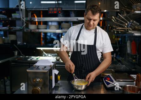 Un chef mûr portant un tablier chuchotant des œufs dans un bol tout en cuisant des pâtes dans la cuisine Banque D'Images