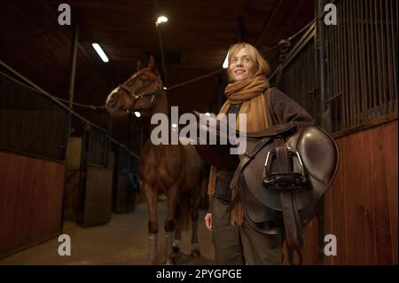 Femme pilote debout et tenant selle à la main au-dessus de son cheval en stable Banque D'Images