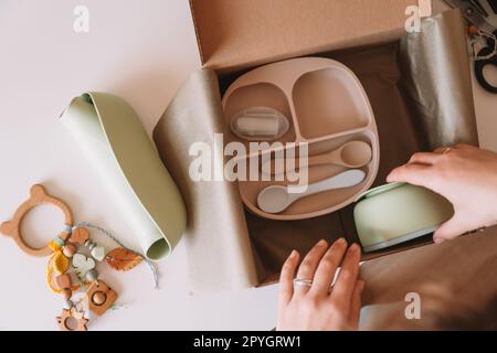 Mains recadrées emballage, emballage de l'expédition avec des plats de bébé en silicone, assiette, cuillères, bol, anneau de dentition écologique dans la boîte. Vue de dessus Banque D'Images