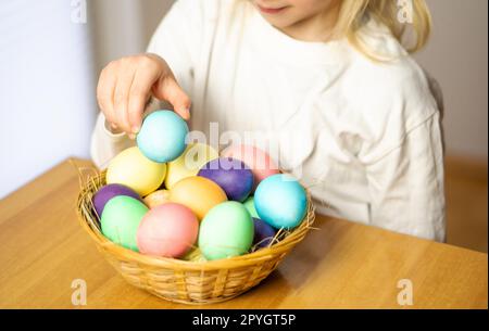 Petite fille blonde, de cinq ans, jouant avec des œufs de Pâques colorés dans un panier sur une table en bois. Banque D'Images