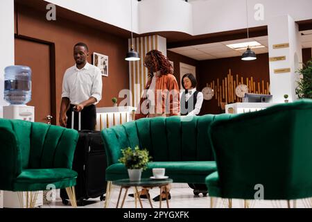 Couple assis dans le salon de l'hôtel, en attente de s'inscrire et de s'enregistrer à l'arrivée. Les jeunes clients arrivant dans un complexe de luxe, discutant avec les employés de la réservation à la réception. Banque D'Images