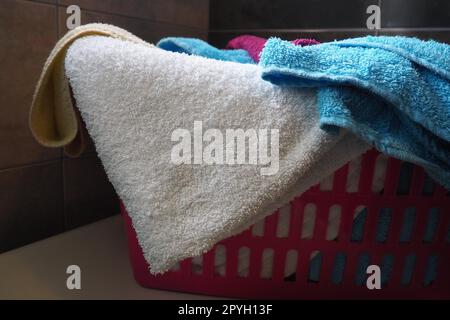 Serviettes dans le panier à linge. Les serviettes en éponge en coton bleu et rose sont jetées dans un panier en plastique rose. Entretien ménager. Stocker et séparer le linge avant du laver. Lumière provenant du haut de la fenêtre ouverte. Banque D'Images