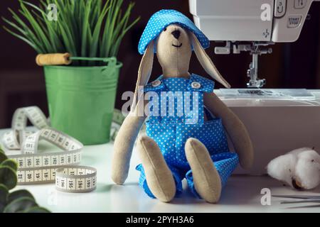 Mignon jouet lapin cousu en costume de design bleu assis près de la plante d'intérieur, ruban à mesurer et coton, penché à la machine à coudre Banque D'Images