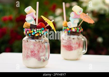 Freakshakes en pots avec paille, guimauve, fraise, chocolat sur une table blanche sur fond de fleurs. Banque D'Images