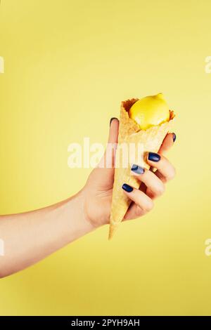 Main féminine avec des ongles bleus tenant le cône de crème glacée gaufrée rempli de citron isolé sur jaune. Vertical. Espace de copie Banque D'Images