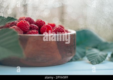 Bol rond en bois partiellement flou plein de framboises juteuses rouges mûres sur fond de bokeh avec des feuilles de framboisier Banque D'Images