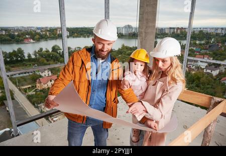 Les parents avec leur fille étudient des dessins architecturaux dans un immeuble en construction. Homme tenant le plan du bâtiment et souriant tout en se tenant à côté de la femme et de la fille sur le chantier de construction. Banque D'Images