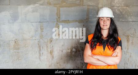 Jeune femme en casque blanc et gilet orange haute visibilité, cheveux longs foncés, mains croisées, en confiance. Rétrogr. Mur site de construction flou Banque D'Images