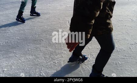 Les enfants montent dans un parc de la ville à la patinoire. Jambes et mains d'un patineur en patinant sur la glace. Patins artistiques habillés sur les jambes. Longues ombres du faible soleil d'hiver Banque D'Images