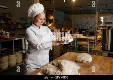 Femme boulangère en uniforme sème la farine à travers le tamis de cuisine sur la pâte Banque D'Images
