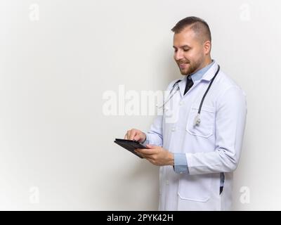 Jeune médecin caucasien avec cheveux courts, moustache et barbe. Habillé en robe blanche et stéthoscope tapant sur l'ordinateur tablette. Tenez-vous devant le mur blanc. Vue latérale Banque D'Images