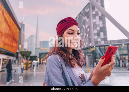 Bonne fille asiatique utilisant le réseau sans fil de smartphone pour se connecter à Internet tout en marchant dans le centre de Dubaï avec la tour Burj Khalifa et d'autres gratte-ciel Banque D'Images