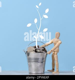 Un mannequin en bois et un seau miniature avec des pièces de monnaie en métal et un arbre peint avec des feuilles sur fond bleu. Concept de croissance des revenus et des bénéfices Banque D'Images