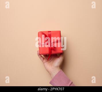 Une main de femme tenant une boîte cadeau rouge enveloppée de ruban sur un fond beige, un concept de félicitations, surprise Banque D'Images
