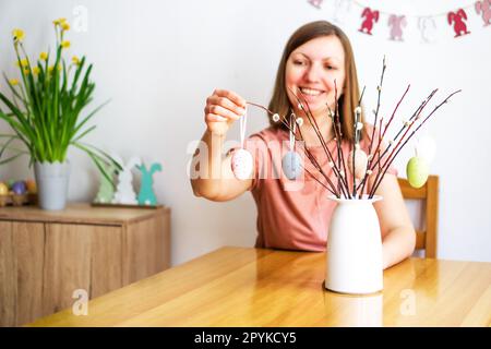 Belle femme souriante décorant un bouquet de branches de saule avec des oeufs de Pâques colorés à la maison. Banque D'Images