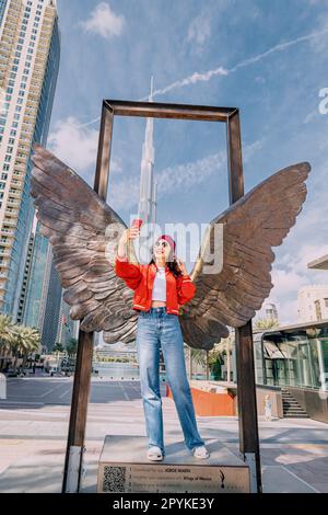 18 janvier 2023, Dubaï, Émirats Arabes Unis : une jeune fille touristique prend des photos de selfie à la statue des ailes du Mexique contre le pittoresque Burj Khalifa et d'autres gratte-ciels Banque D'Images