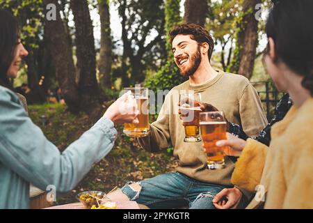 Un homme caucasien barbu et une femme caucasienne à long poil toast avec des tasses de bière dans un pub de forêt en plein air, entouré de personnes méconnaissables, tous smil Banque D'Images