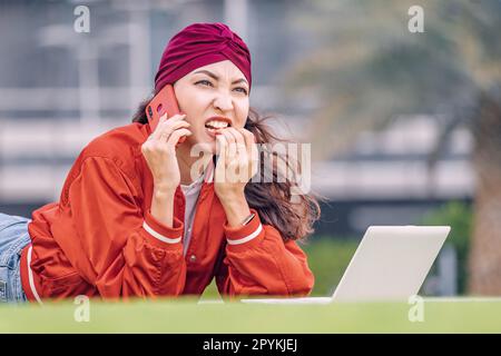 Une femme indienne frustrée et effeurée qui mord ses ongles parle par téléphone et travaille à l'extérieur sur son PC portable Banque D'Images