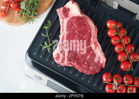 Steaks de première qualité crus sur le gril électrique. Steaks de viande de choix black angus Ribeye sur un grill électrique moderne au romarin, à la tomate cerise et au poivre. Cuisine pro Banque D'Images