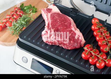 Steaks de première qualité crus sur le gril électrique. Steaks de viande de choix black angus Ribeye sur un grill électrique moderne au romarin, à la tomate cerise et au poivre. Cuisine pro Banque D'Images