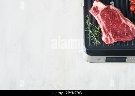 Steaks de première qualité crus sur le gril électrique. Steaks de viande de choix black angus Ribeye sur un grill électrique moderne au romarin, à la tomate cerise et au poivre. Cuisine pro Banque D'Images