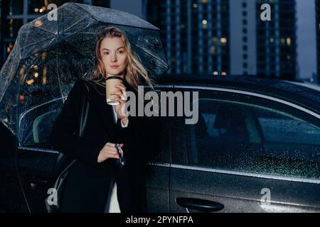 Une femme se tient près de sa voiture lors d'une soirée pluvieuse sous un parapluie. Boire du café à emporter Banque D'Images