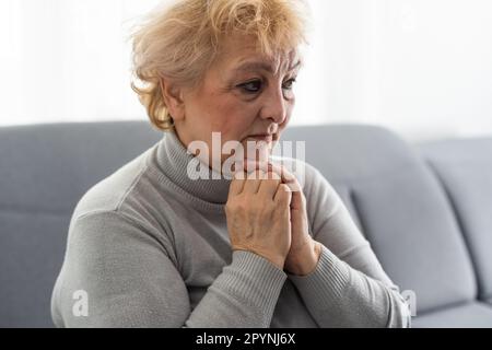 Frowning bouleversé femme mature mettre la tête sur les mains pliées, en regardant dans la distance. Perdu dans les pensées bouleverser la femme assise seule, tête tourné gros plan portrait Banque D'Images
