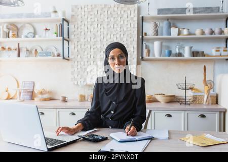 Jeune femme prospère et confiante en hijab noir et burqa assis à la maison à la table de cuisine et travaillant à distance avec des documents et un ordinateur portable. Il regarde Banque D'Images