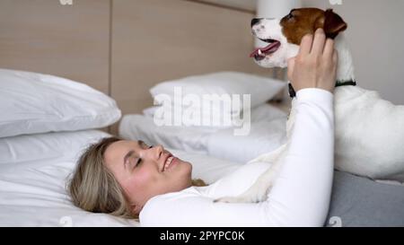 Une belle fille se trouve sur le lit, et sur elle se trouve le chien Jack Russell. La fille dort avec son chien au lit. Jack Russell Terrier liche son propriétaire Banque D'Images