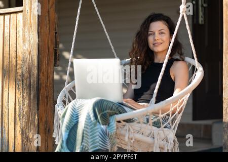 une jeune femme se repose sur une chaise de balançoire macrame avec ordinateur portable sur la terrasse de la maison de campagne