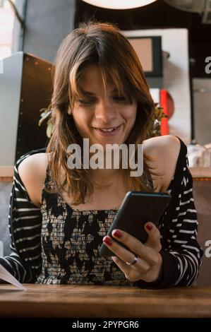 portrait d'une jeune femme transgenre latine d'origine argentine, dans un restaurant assis au téléphone et souriant, image verticale. Banque D'Images