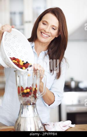 Dans l'attente d'une matinée fructueuse. Une femme attirante qui ajoute des fruits à un mélangeur tout en se tenant sur un comptoir de cuisine. Banque D'Images