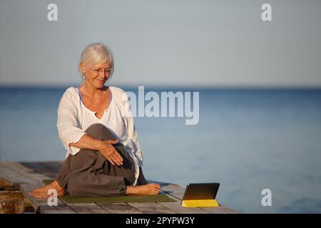 S'assurer qu'elle obtient les postures juste. Une femme mûre faisant des exercices de yoga sur une jetée avec l'aide de sa tablette numérique. Banque D'Images