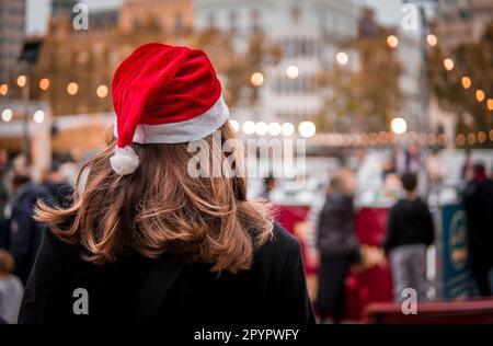 Photo de la femme Brunette vue de derrière avec chapeau du Père Noël regardant les gens patiner sur glace dans un carré avec des lumières de Noël Banque D'Images