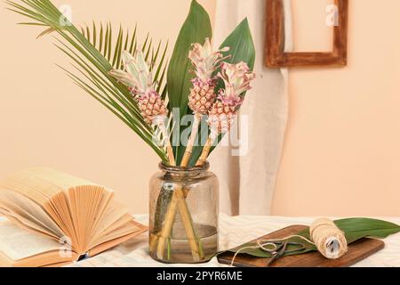 Vase avec ananas décoratif, feuilles de palmier, livre et corde sur la table près du mur beige Banque D'Images