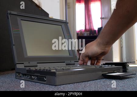 mains d'une personne prenant une disquette et la plaçant dans un vieil ordinateur portable. La personne est sur le sol d'une pièce Banque D'Images