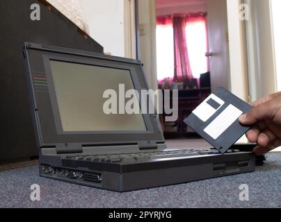 mains d'une personne prenant une disquette et la plaçant dans un vieil ordinateur portable. La personne est sur le sol d'une pièce Banque D'Images